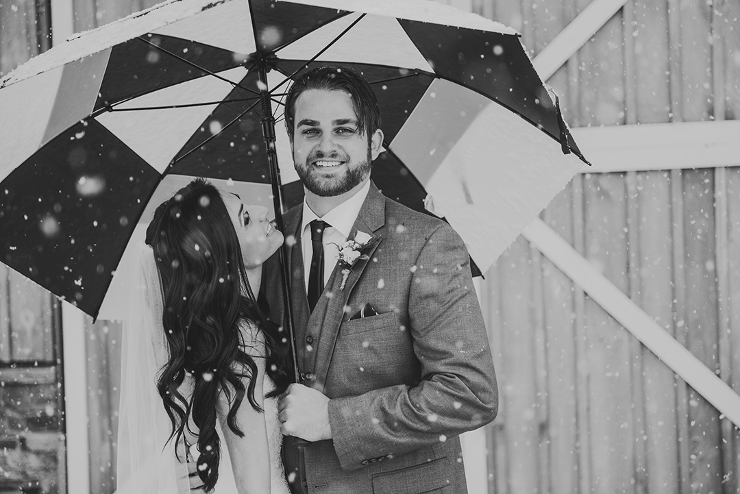 a groom smiles while his bride looks at him in a November snowstorm