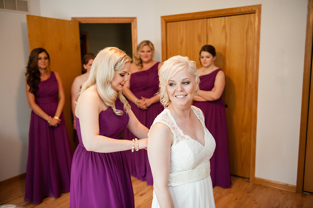a made of honor buttoning brides dress as the bridesmaids watch