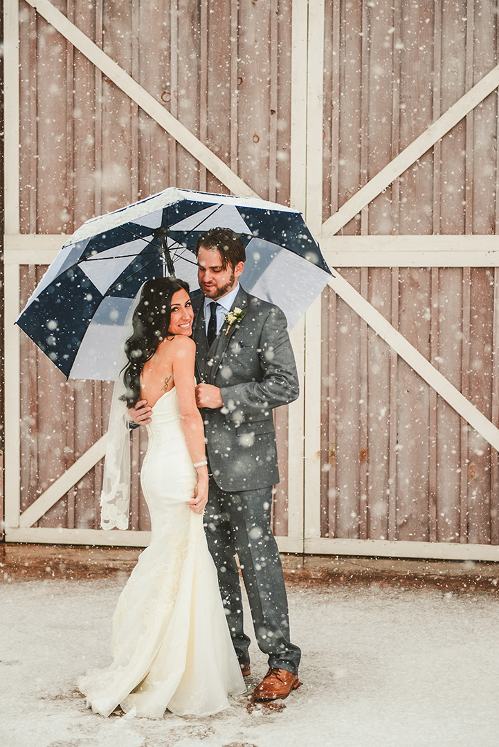 a bride shows off the back of her dress in front of a rustic barn