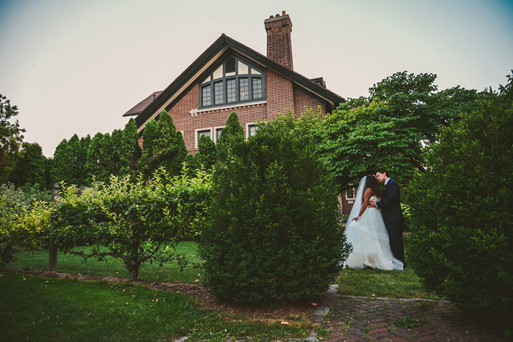 a husband and wife sneaking a kiss between two bushes at the Chaney Mansion