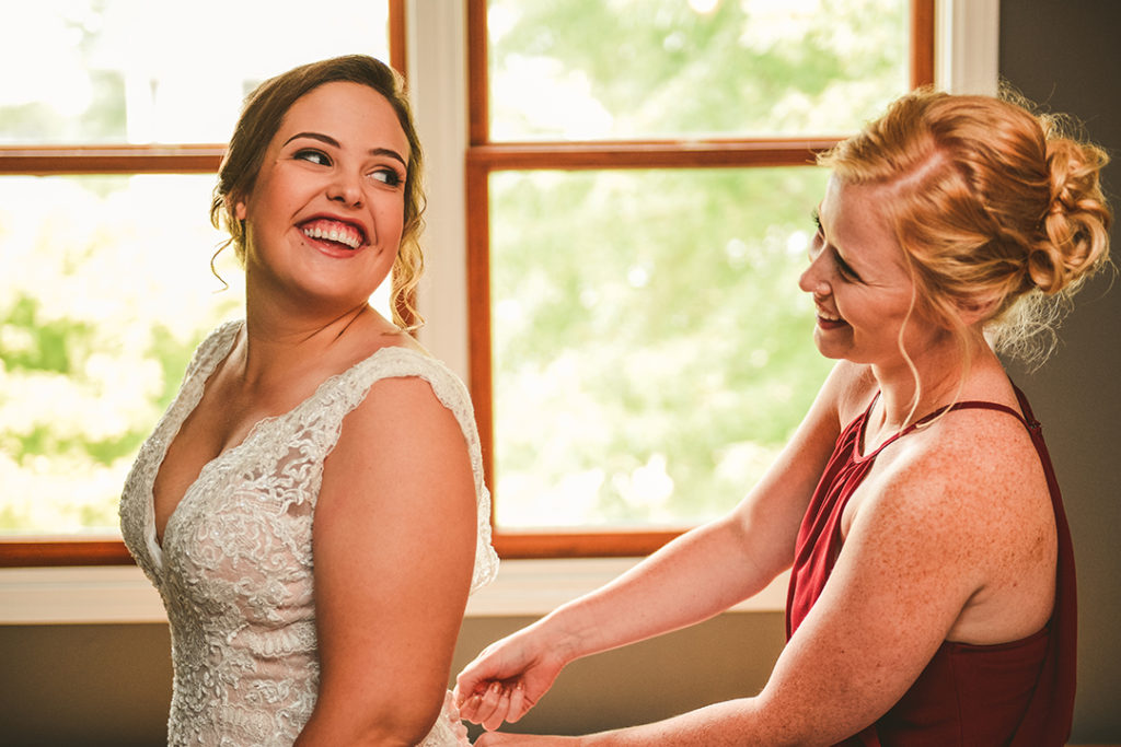a bride and her maid of honor laughing as the bride gets her dress on
