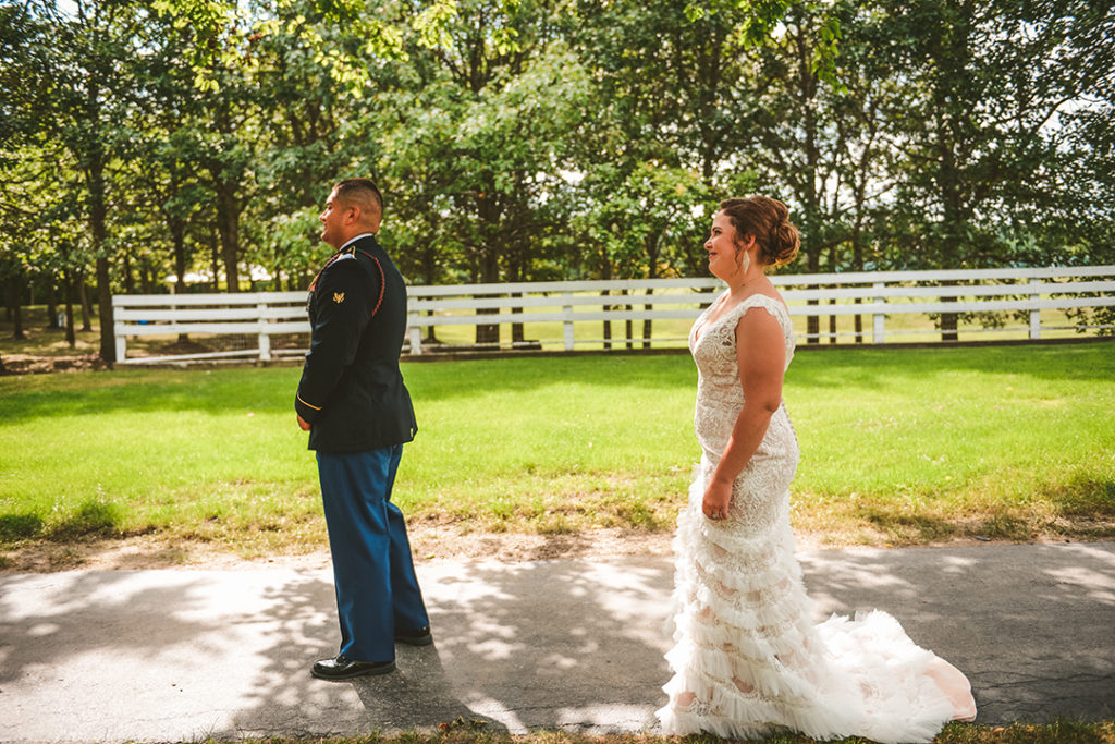 the bride walking up behind her groom during the first look by a white farm fence