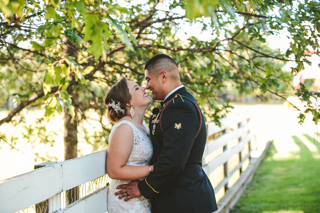 newly weds sneaking a kiss on a old white fence with trees surrounding them