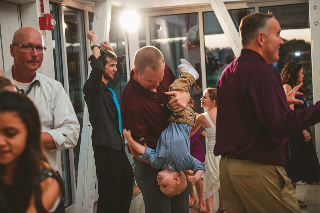 a man holding a boy upside down at a wedding reception with people dancing in the background