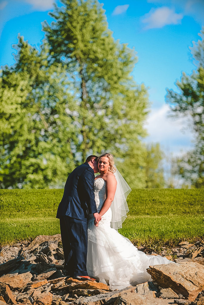 a husband kissing his wives neck with a brilliant blue sky and trees in the background