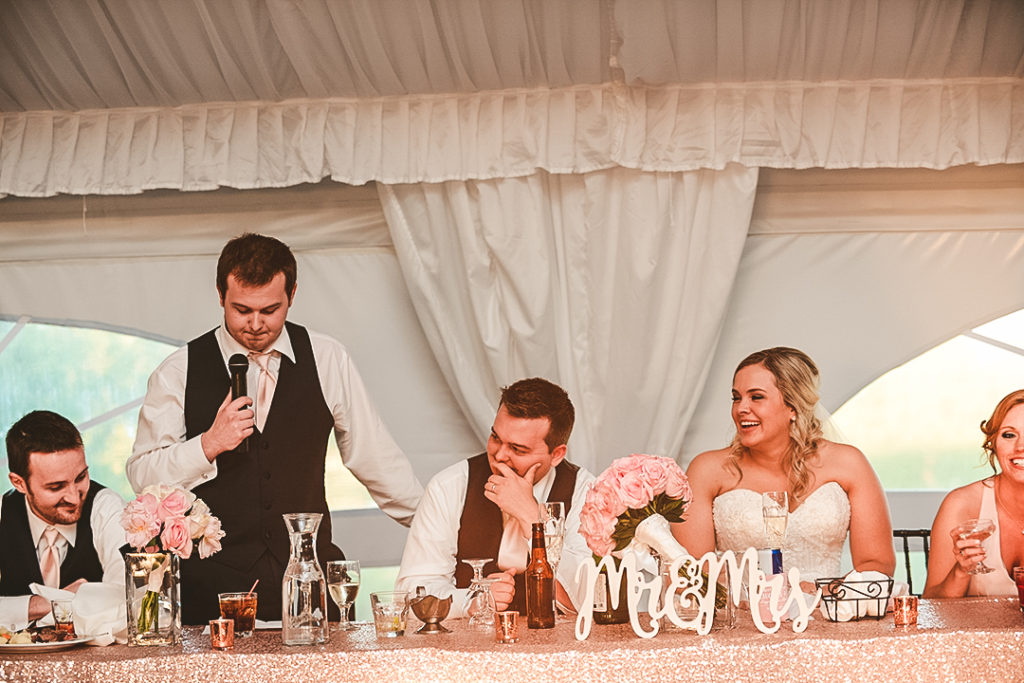 a bride and groom laughing at the head table as the best man gives his speech