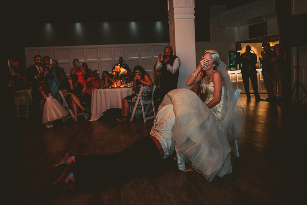 bride laughing as her groom takes off her garter at a wedding reception