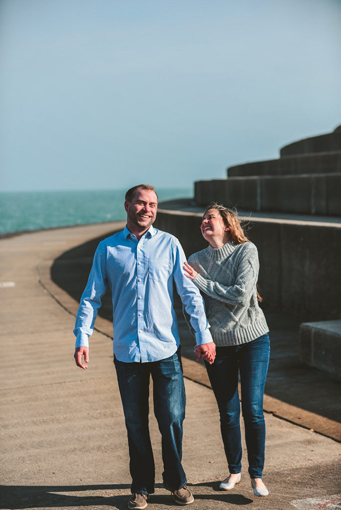 a man and a woman laughing as they walk down the lakefront in front of blue water