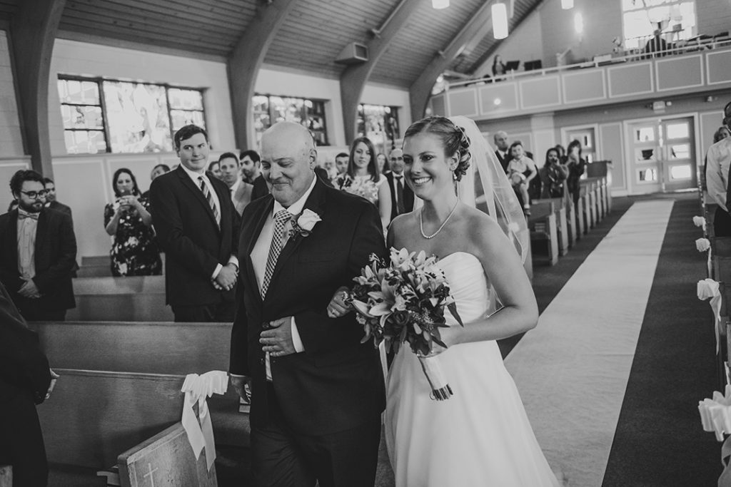 a father walking his daughter down a church aisle as the bride smiles