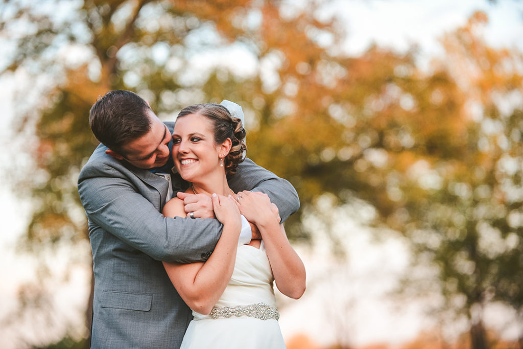 a man hugging his wife from behind as she looks back and smiles in the fall