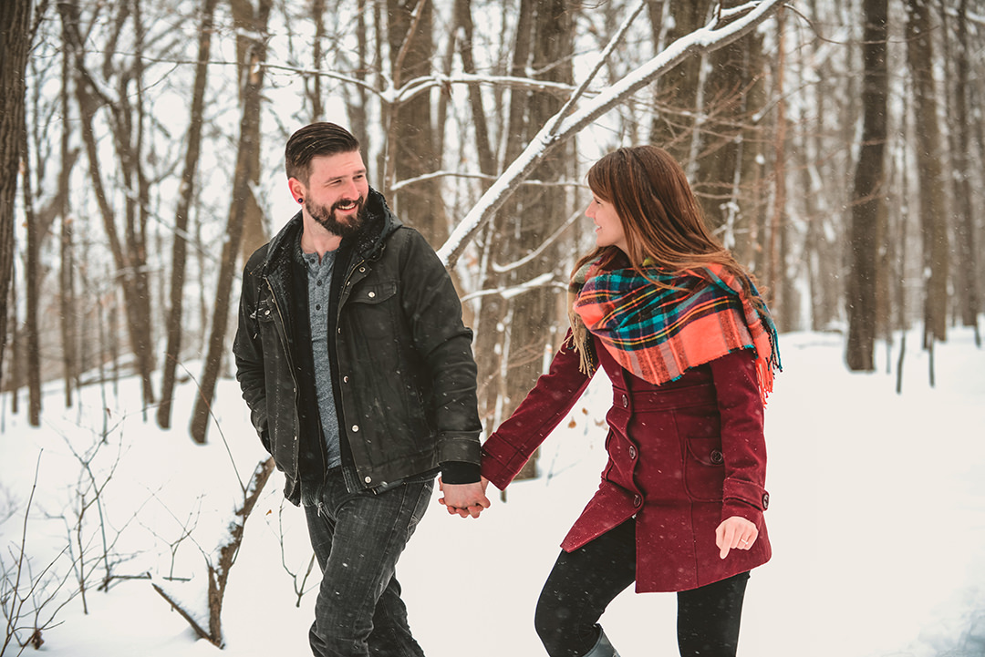 a engaged couple holding hands as they walk in a snowy woods in Shorewood