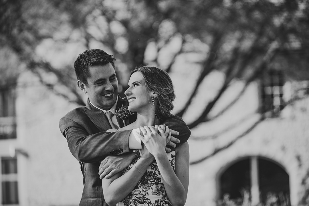 B & W of a groom hugging his wife from behind in front of an old growth tree