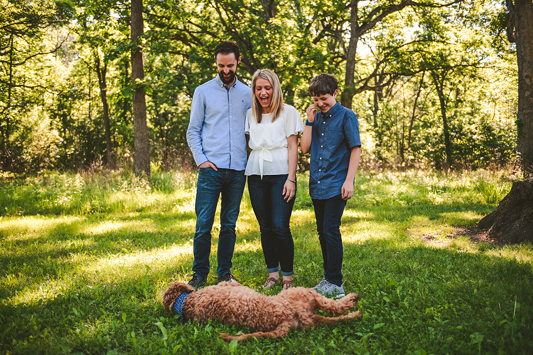 family laughing as their dog rolls in grass on a family photo session