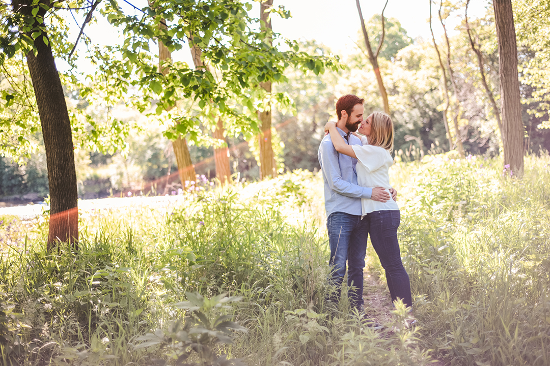 a man and a woman kissing with their arms around each other with a lens flare in the woods