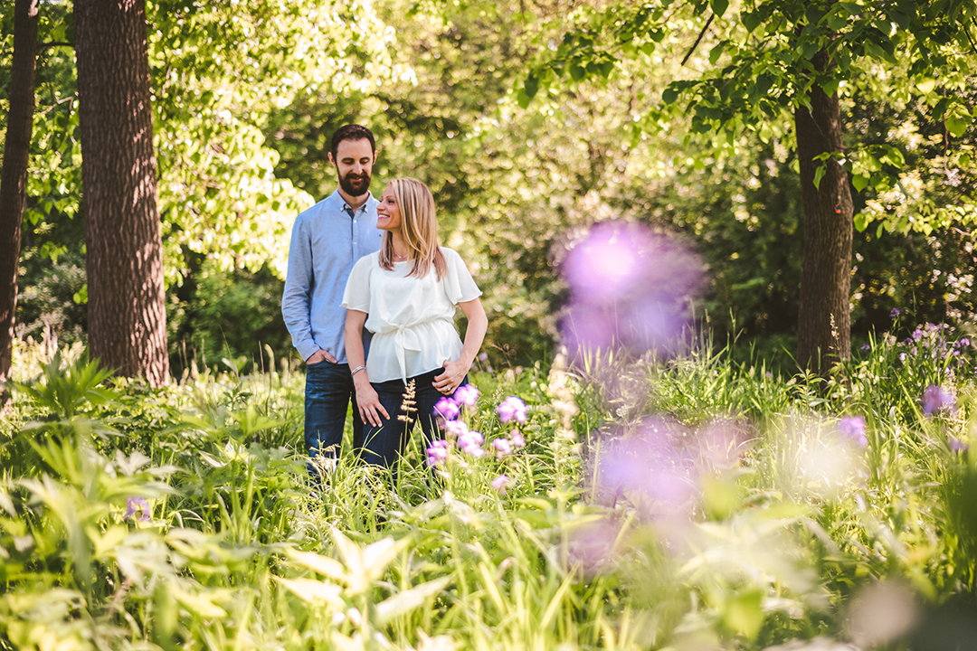 a woman looking away as her boyfriend looks at her in the woods with purple flowers