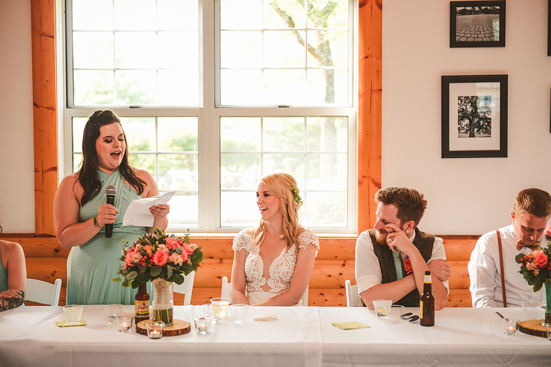 the bride and groom laughing as the maid of honor gives her speech in front of a bright window