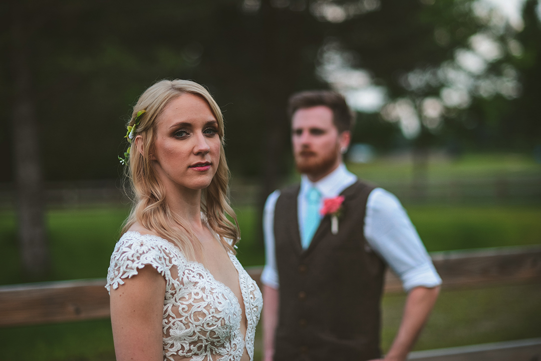 a bride looking towards the camera as her groom stands in the background as the sun sets