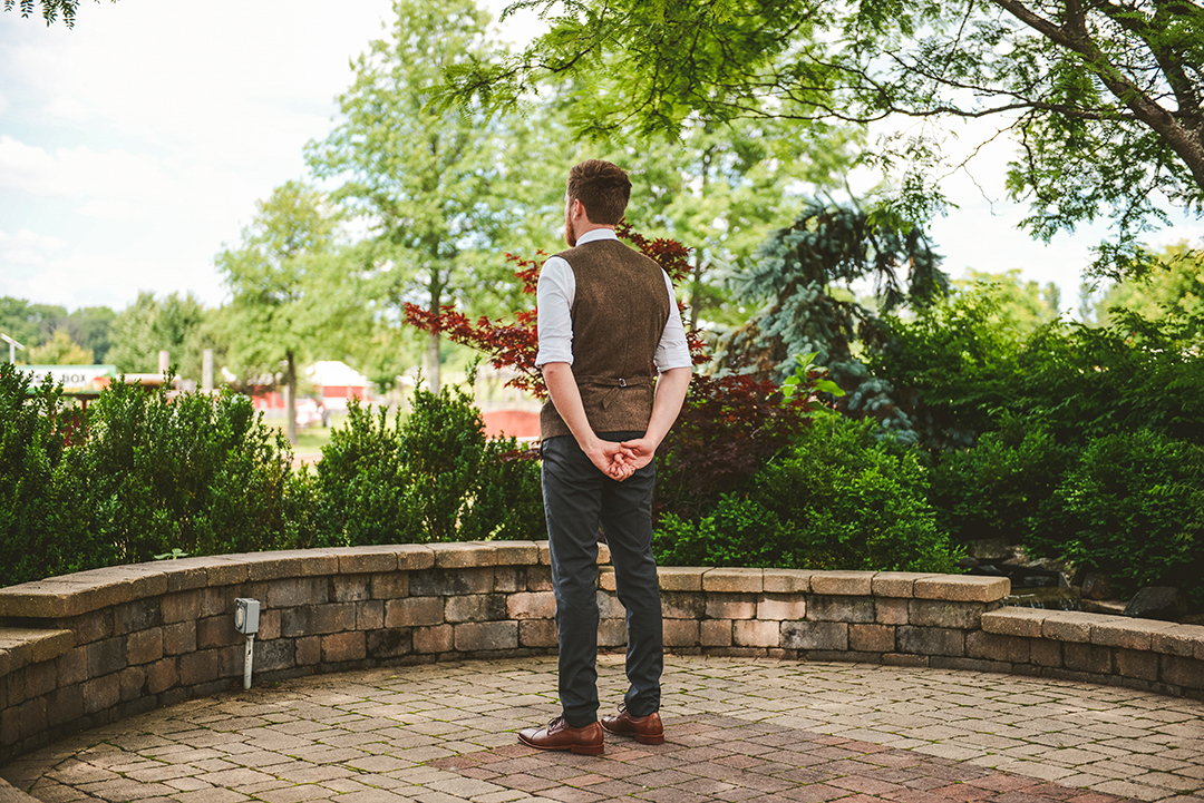 a groom standing with his arms behind his back as he waits for his bride to come up behind him for their first look