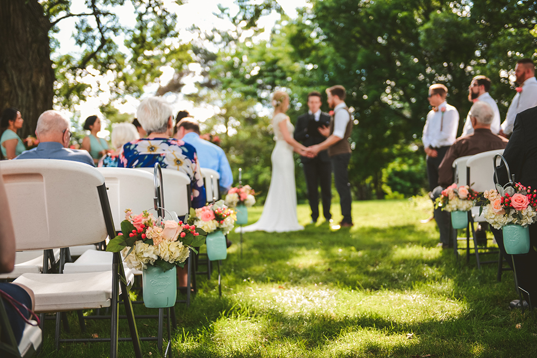 a detail of flowers at an outdoor wedding with the bride and groom holding hands in the background