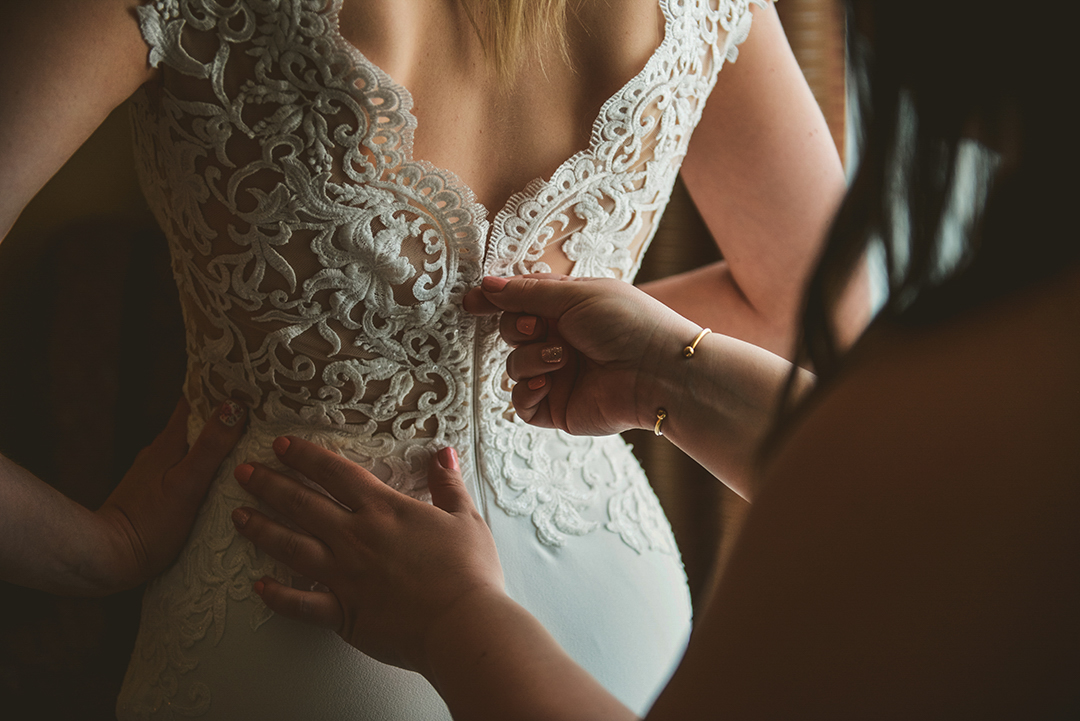 a detail of a bridesmaid zipping up the brides dress as the bride stands with her hand on her hip