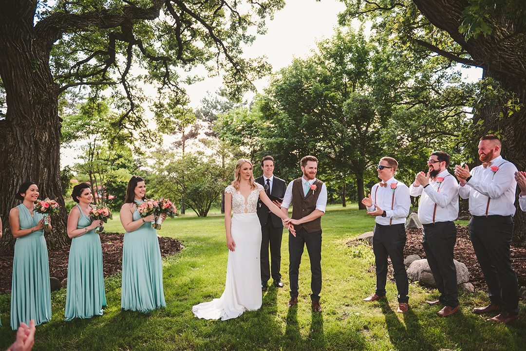 a bride and groom laughing with their bridal party under two large trees at a apple orchard