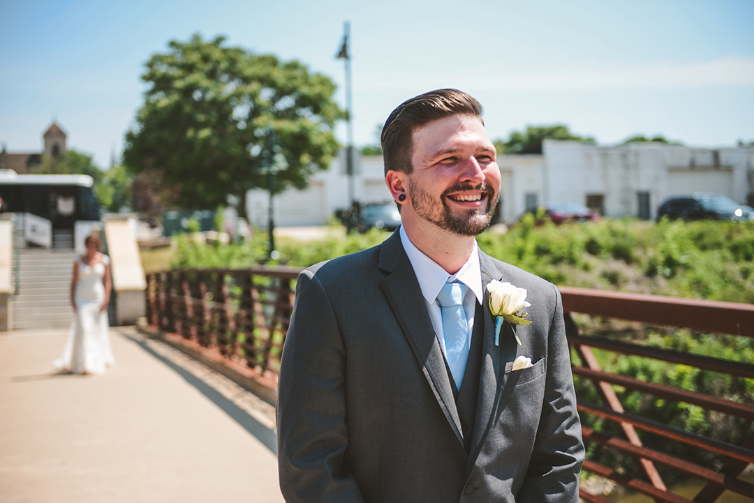 the groom smiling as his bride walks up behind him at a bridge in Lemont