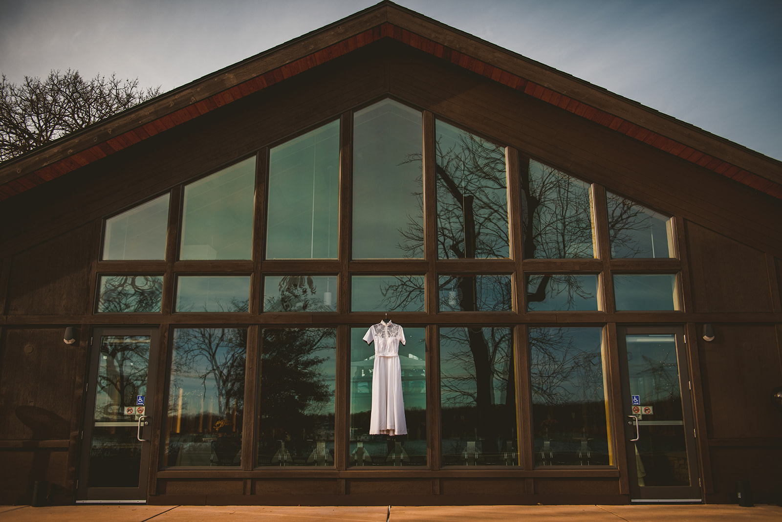 A vintage wedding dress hanging in front of a large bank of windows on a fall evening