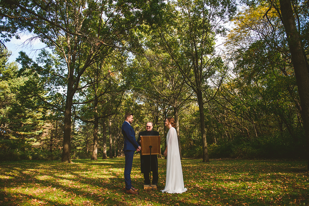 a wide image of a bride and groom getting married in front of old growth trees in the fall at the Saint James Farm in Warrenville IL