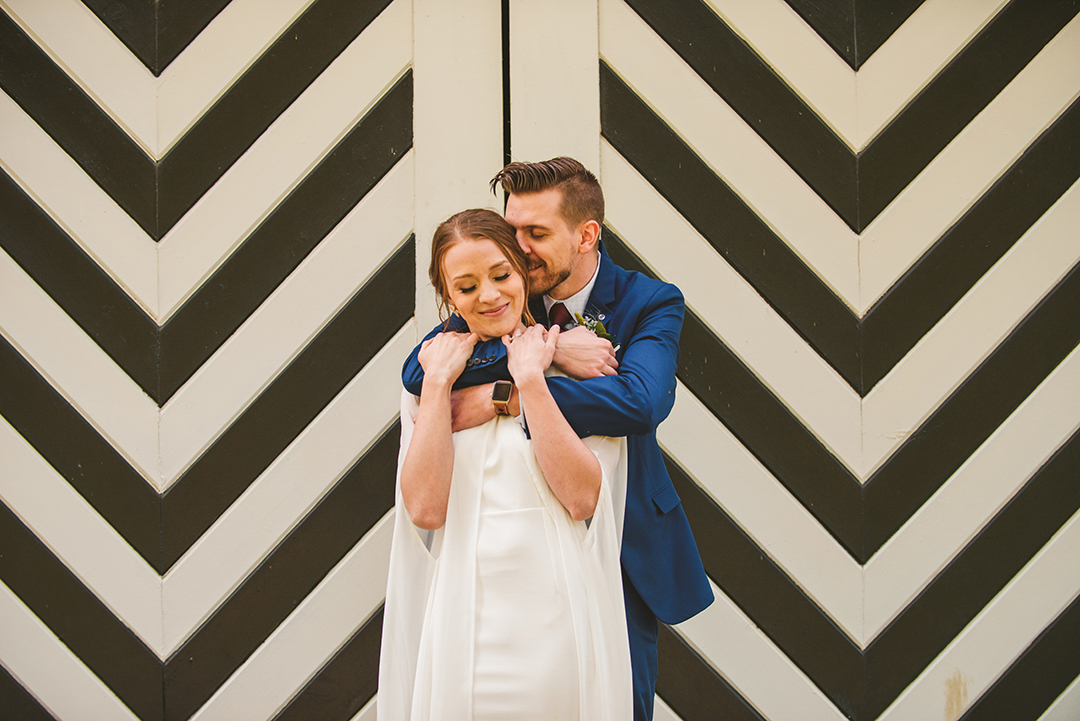 a groom kissing his brides head with his arms around her in front of a black and white striped door at the Saint James Farm in Warrenville IL