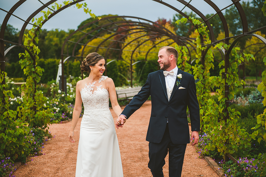 a bride and groom holding hands as they walk down a beautifully planted path at a Cantigny Park wedding in the fall