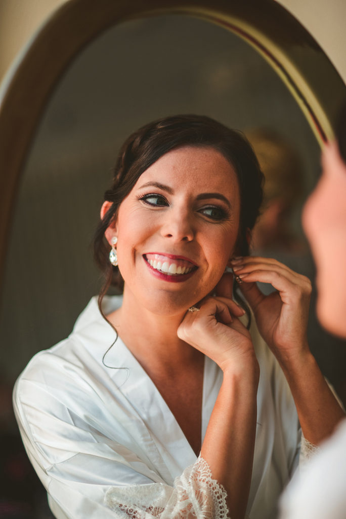 the bride smiling into a mirror as she puts on her earrings
