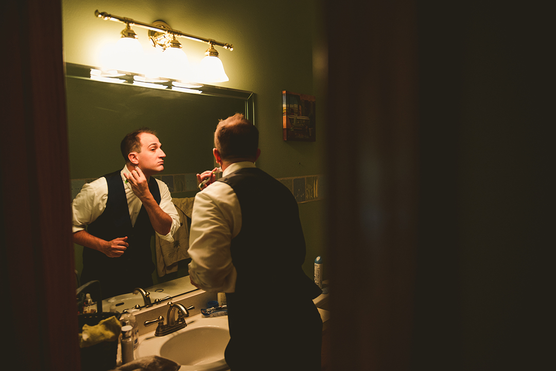 the groom shaving in a dimly lit bathroom before he leaves to get married
