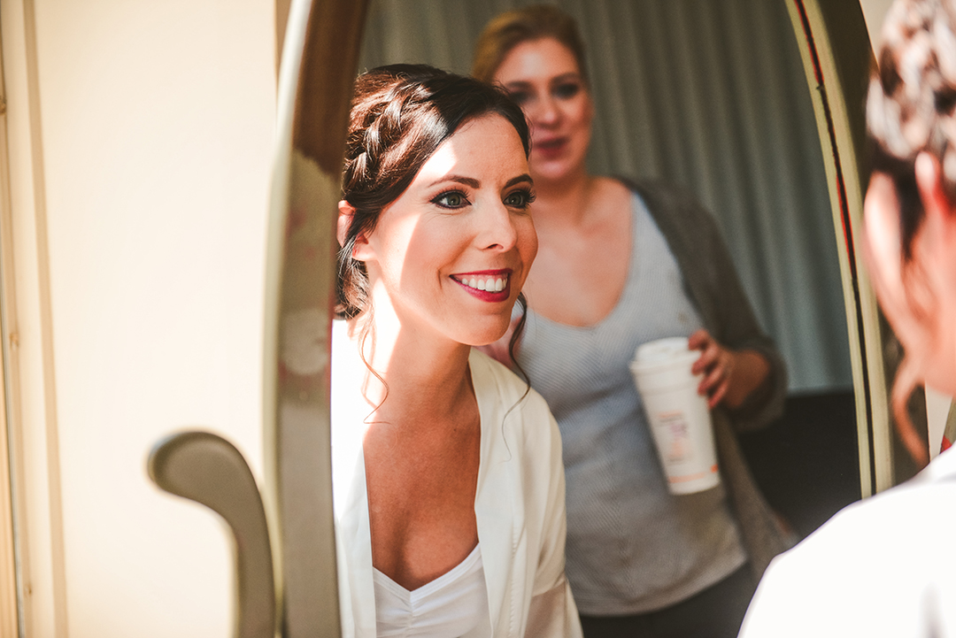 a bride looking at herself in a mirror smiling as the makeup artist watches