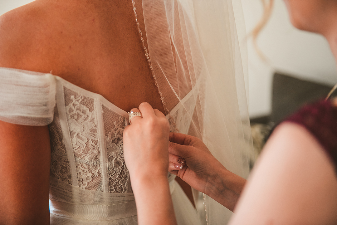 a detail of the maid of honor zipping up the brides dress in a warmly lit room