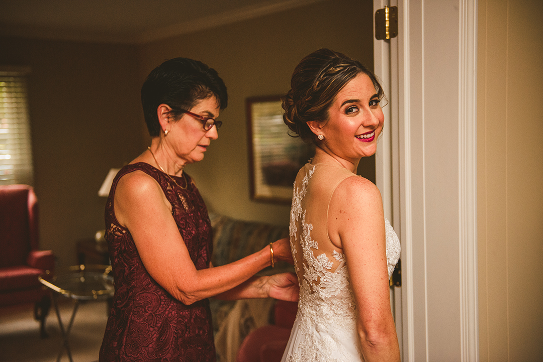 the mother of the bride zipping up the brides dress as she smiles right before her wedding at St Thomas Apostle Church in Naperville