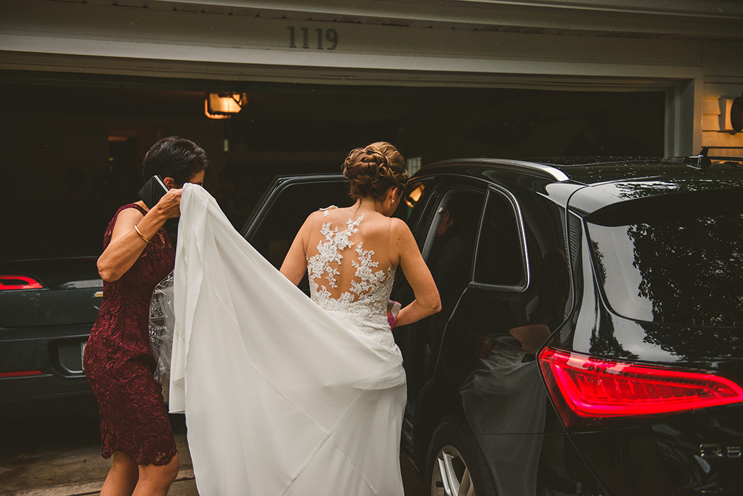 the mother of the bride holding the train of the brides dress as she gets into her car to head to her wedding at St Thomas Apostle Church in Naperville