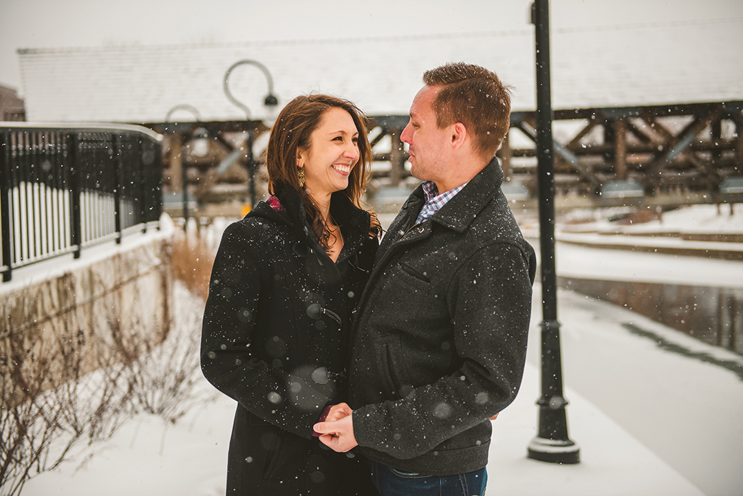 a bride and groom holding hands as they laugh in the snow on the downtown Naperville riverwalk