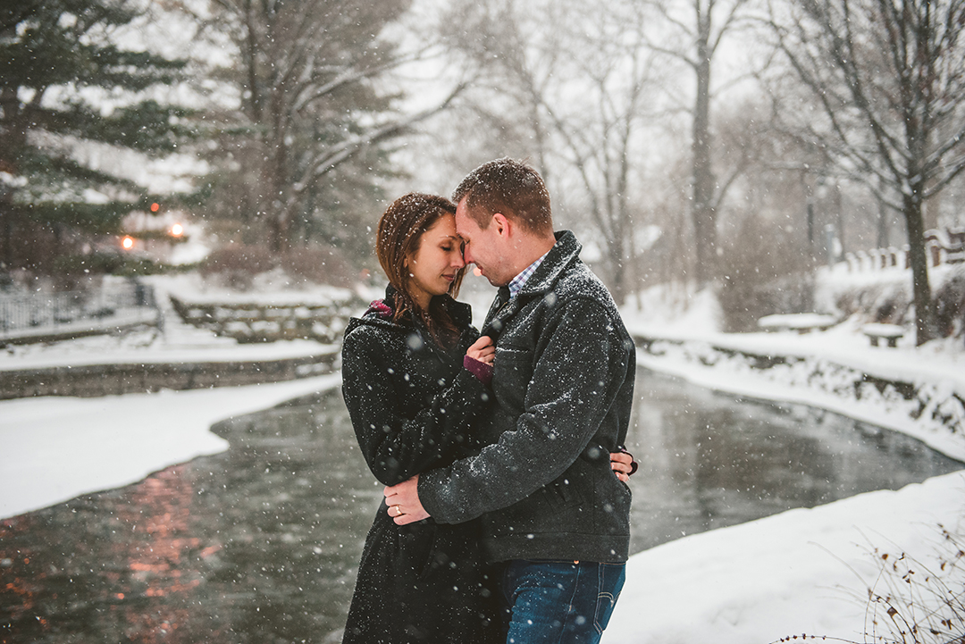 a groom and bride with their heads together as the snow falls down on them on the Naperville Riverwalk 