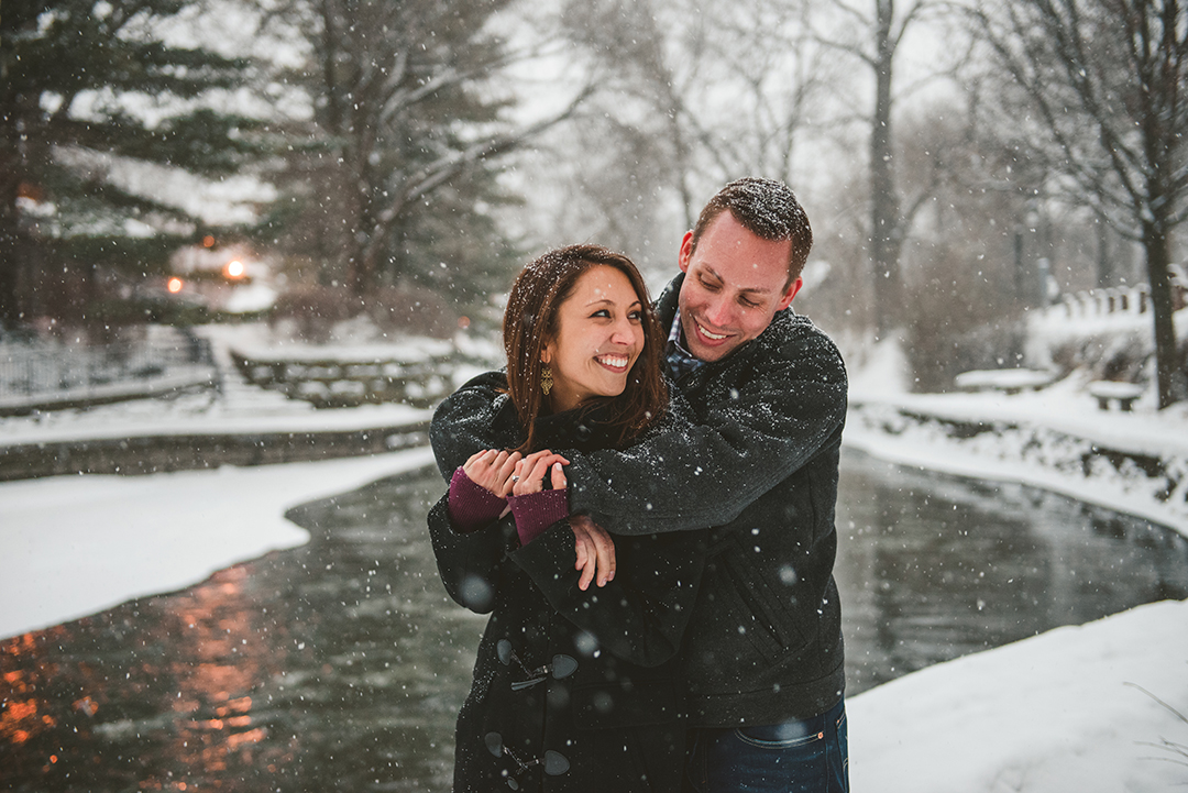 A groom hugging his bride from behind as they laugh in the snow on the Naperville Riverwalk in winter