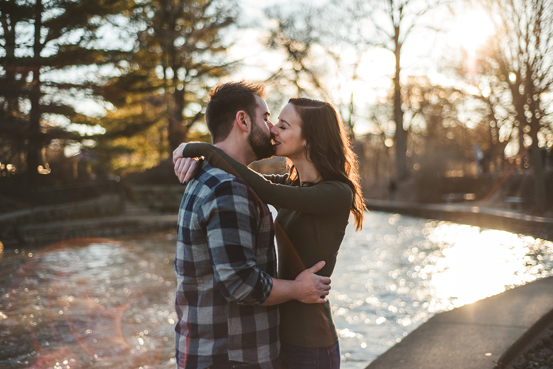 a man and a woman kissing as they smile by a river with the setting sun while capturing Naperville Riverwalk engagement photography in the spring