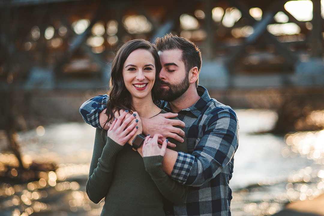 a man holding onto his fiance as she smiles and hugs into him during their engagement session on the Naperville Riverwalk