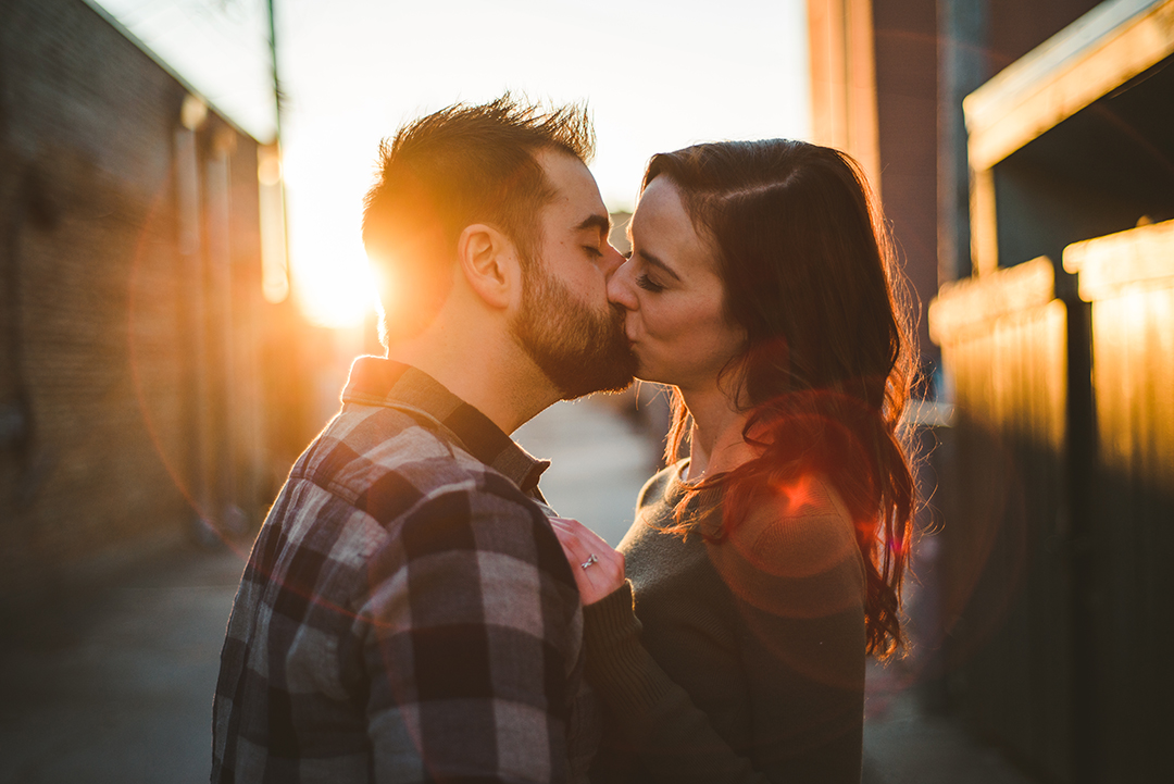 a man and a woman kissing as the sun sets in the background by the Naperville Riverwalk with a strong sun flare across the frame