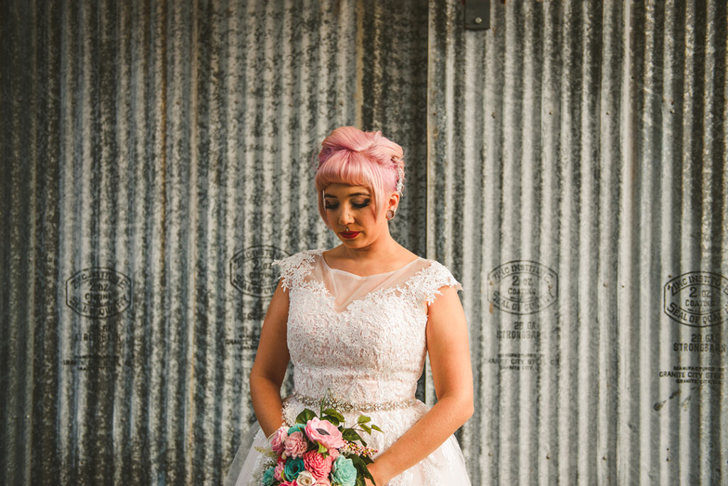 a tattooed bride looking down at her flowers in front of rustic metal at the Warehouse 109