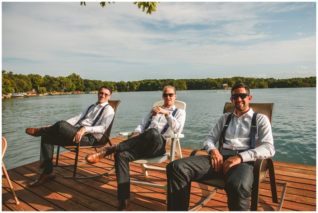 three groomsmen sitting on a dock at a lake wedding right before it starts in the summer at Lake Bloomington