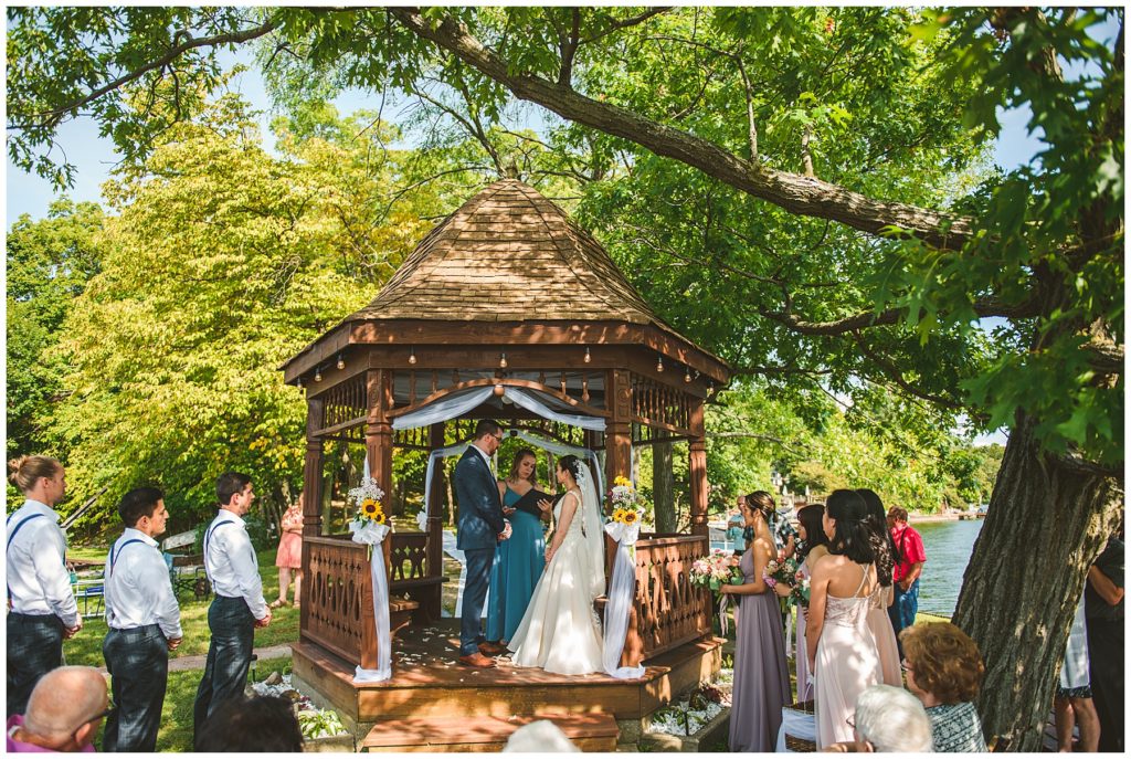 a bride and groom standing wit their bridal party at a handmade wooden gazebo at a gorgeous summer lake wedding