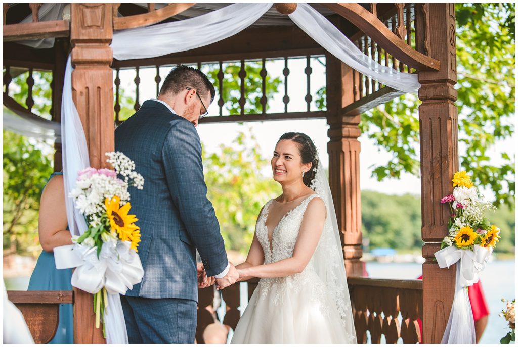 a bride laughing during her vows as her groom holds her hands at their summer lake wedding
