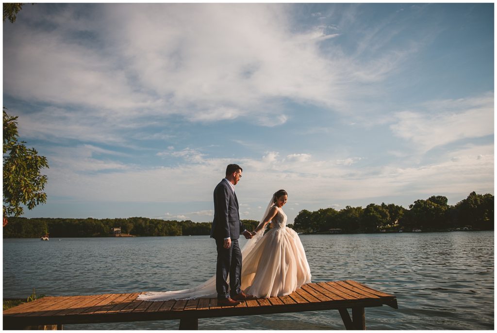 a bride leading a groom down a dock at their summer lake wedding at Lake Bloomington with wispy clouds in the background