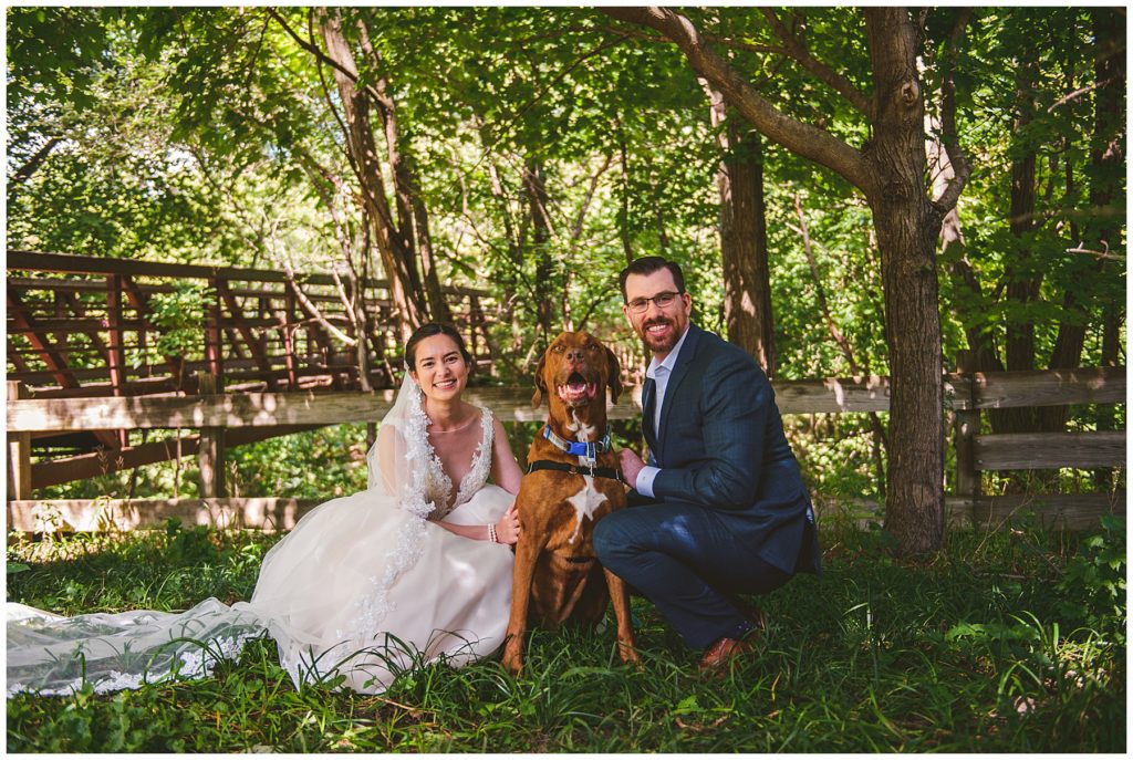 a bride and groom standing with their dog by a wooden bridge at their summer lake wedding