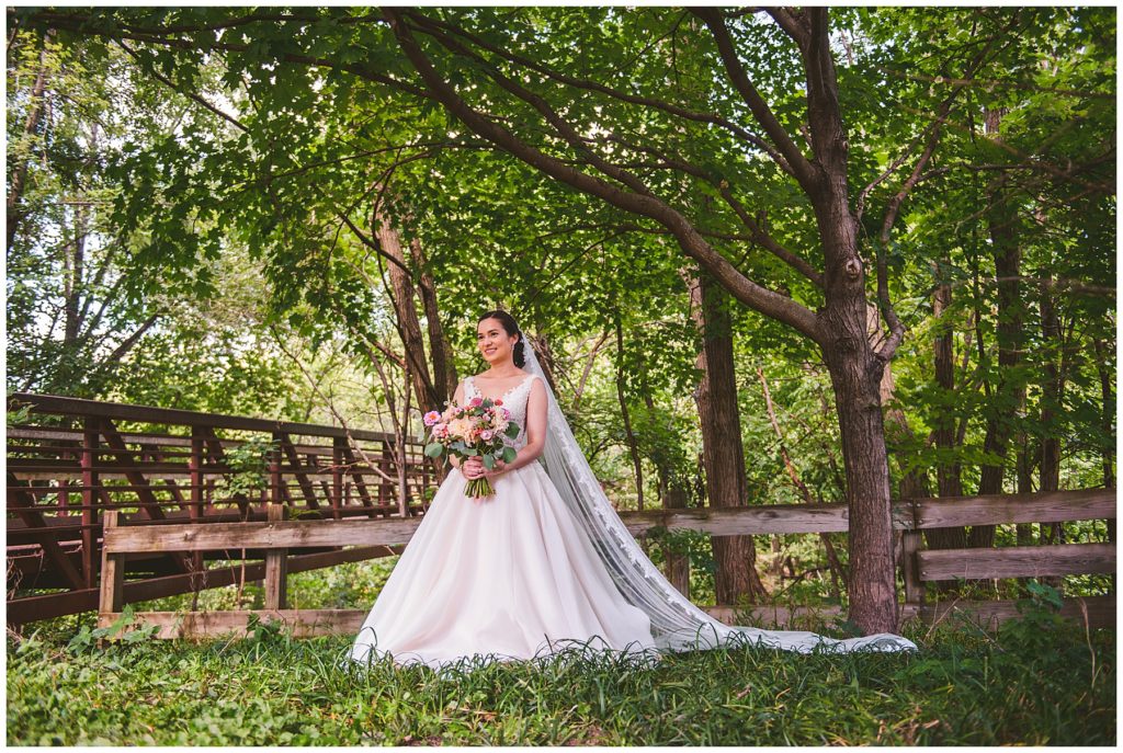 a breathtaking bride standing with her vail spread out in front of a vintage bridge at her summer lake wedding