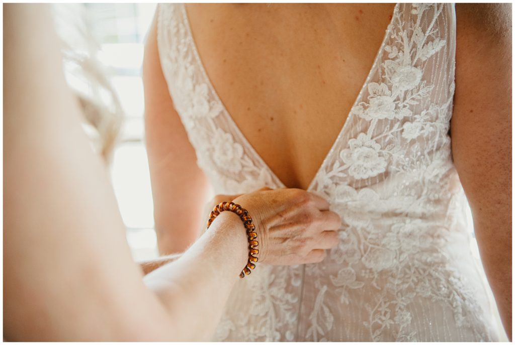 a close up of the mother of the bride buttoning up the back of the brides dress on her wedding day